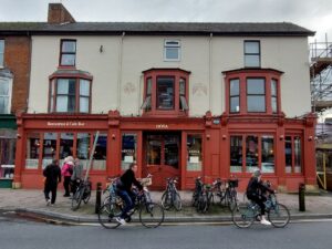 Photo of Dosa Darlings restaurant in Cowley Road, with a full cycle rack in front, pedestrians, and two people cycling past.