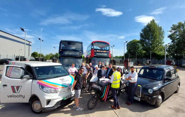 Transport Modes Unite To Support Congestion Charge A photo of a group of people in front of an electric van, a black taxi, a CoWheels car cub car, two buses a cargo bike an electric scooter and another bicycle.