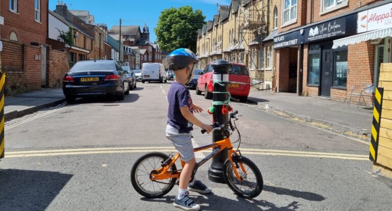 A small child on a bicycle looking down a street made safe with a bollard and planters. The bollard is decorated with fuzzy baubles and he is reaching out to play with them.